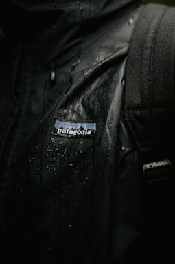 Close-up of charcoal streetwear jacket with water droplets, styled against a wet urban pavement backdrop.