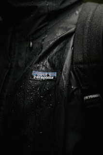 A close-up view of a black jacket with visible water droplets on its surface, featuring a Patagonia logo. The texture of the fabric and the wet sheen are clearly visible.