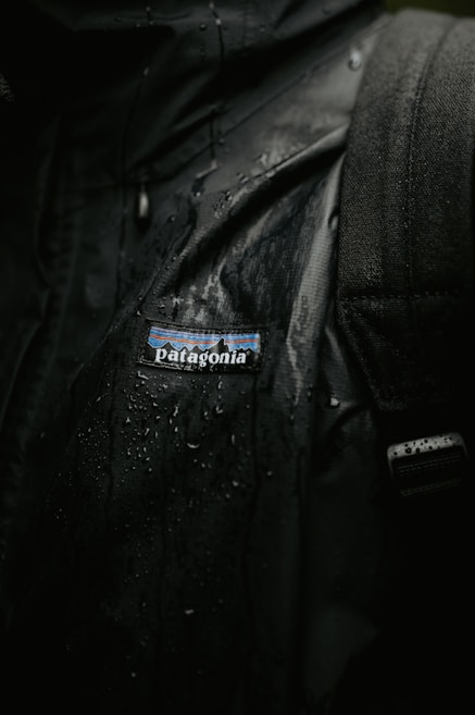 A close-up view of a black jacket with visible water droplets on its surface, featuring a Patagonia logo. The texture of the fabric and the wet sheen are clearly visible.