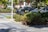 A landscaped urban area with a pedestrian walkway lined with textured paving stones, surrounded by lush greenery and tropical plants. Two cars are parked on the street, one of which is identifiable as a Street View vehicle with a camera mounted on top. The scene includes a streetlight pole and some buildings in the background.