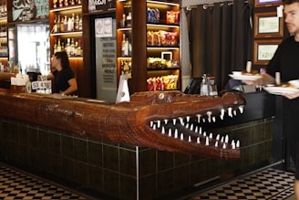 A bar interior featuring a large wooden crocodile sculpture integrated into the bar counter. Behind the counter, bottles of various beverages are displayed on illuminated shelves. A person is serving drinks, and another is carrying plates of food.