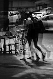 A black and white photograph features a reflective scene with overlapping images of a person walking, cars on the street, and a bicycle. The reflection in the glass creates a layered effect, blending various elements together.