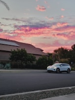 A white electric car is parked on a quiet suburban street. The sky above is vibrant with shades of pink, purple, and orange, indicating a colorful sunset. The foreground features a traditional suburban house with a tiled roof and solar panels. Lush, green trees and shrubs accentuate the serene atmosphere.