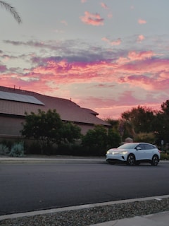 A white electric car is parked on a quiet suburban street. The sky above is vibrant with shades of pink, purple, and orange, indicating a colorful sunset. The foreground features a traditional suburban house with a tiled roof and solar panels. Lush, green trees and shrubs accentuate the serene atmosphere.