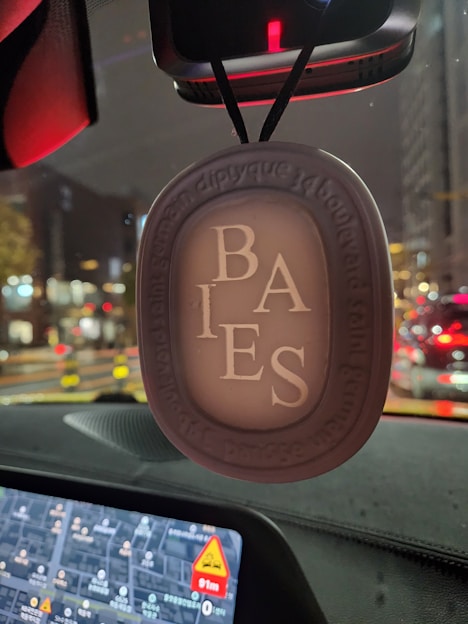An air freshener labeled 'BAIES' hangs from a rearview mirror in a car. The background shows a blurry view of a city street at night, with colorful lights and vehicles. The dashboard features a digital map with visible directions.