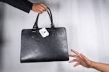 A black leather handbag is being passed from one hand to another. The bag has a white tag attached to it, displaying a logo and the word 'Obsidian'. The background is a simple light gray, highlighting the focus on the handbag and hands.