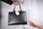 A black leather handbag is being passed from one hand to another. The bag has a white tag attached to it, displaying a logo and the word 'Obsidian'. The background is a simple light gray, highlighting the focus on the handbag and hands.