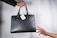 A black leather handbag is being passed from one hand to another. The bag has a white tag attached to it, displaying a logo and the word 'Obsidian'. The background is a simple light gray, highlighting the focus on the handbag and hands.