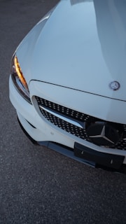 A close-up view of the front section of a white Mercedes-Benz car. The focus is on the grille and the emblem, with a clear view of one headlight emitting an amber light. The surface is shiny, reflecting the surrounding light.