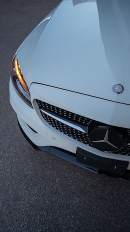 A close-up view of the front section of a white Mercedes-Benz car. The focus is on the grille and the emblem, with a clear view of one headlight emitting an amber light. The surface is shiny, reflecting the surrounding light.