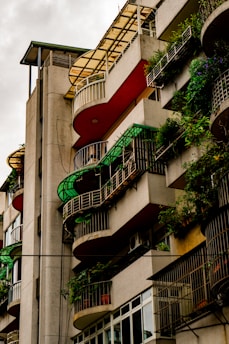 A multi-story building features curved balconies adorned with green plants. The architecture includes metal railings and a mix of red and beige colors, blending urban design with natural elements.