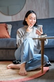 A smiling woman using the myangel app on her phone while sitting in a cozy living room.