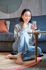 Happy young woman happily using her phone to apply for an online loan in a cozy modern living room.