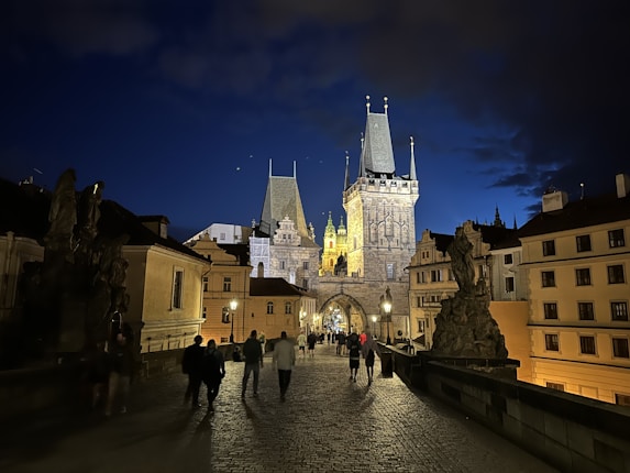 A historic bridge with cobblestone pavement is illuminated by streetlights, with several figures walking towards an ornate, well-lit Gothic-style tower. Statues are visible on either side of the path, and the sky is a deep twilight blue.
