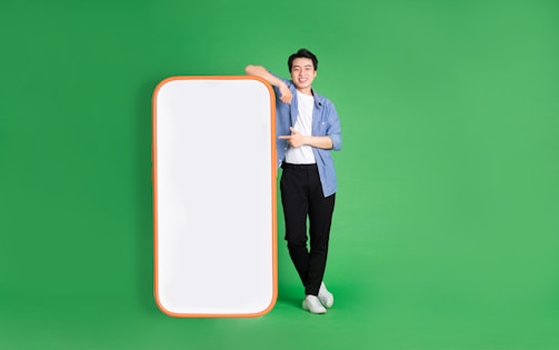 A person stands confidently against a bright green background, leaning on a large, blank smartphone cutout with a red-orange border. They are casually dressed in a blue shirt, white t-shirt, black pants, and white sneakers, and display a positive demeanor.