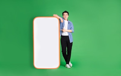 A person stands confidently against a bright green background, leaning on a large, blank smartphone cutout with a red-orange border. They are casually dressed in a blue shirt, white t-shirt, black pants, and white sneakers, and display a positive demeanor.