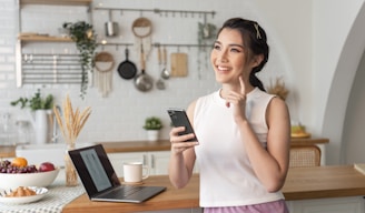 A woman is standing in a modern kitchen, smiling and holding a smartphone. She is wearing a white sleeveless top and pink pants. On the wooden table in front of her, there is an open laptop, a white mug, a bowl of fruit, and some bread. The background features white subway tile walls with various kitchen utensils hanging from a rod, along with some green plants for decoration.