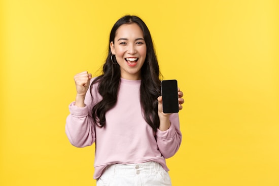 A person with long black hair, wearing a pink sweatshirt and white pants, is joyfully holding up a smartphone with one hand and expressing excitement with a clenched fist. The background is a bright yellow.