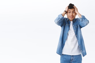 A person wearing a denim shirt and jeans is holding their head with a surprised or anxious expression. The background is plain and white.