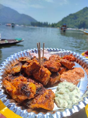 A plate of grilled, spiced kebabs garnished with wooden skewers sits on a reflective silver dish. Accompanying the kebabs are two dollops of sauce, one reddish and the other creamy white with herbs. In the background is a scenic lake surrounded by green mountains, with several boats floating on the water under a clear blue sky.