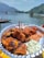 A plate of grilled, spiced kebabs garnished with wooden skewers sits on a reflective silver dish. Accompanying the kebabs are two dollops of sauce, one reddish and the other creamy white with herbs. In the background is a scenic lake surrounded by green mountains, with several boats floating on the water under a clear blue sky.