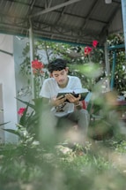 A young person sits outdoors reading a book amidst greenery and flowers. The metal roof structure provides shade, while vibrant red flowers add color to the surroundings.