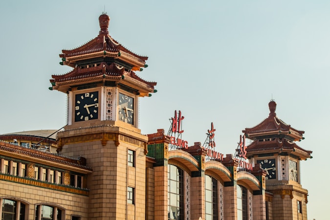 A traditional Chinese architectural structure featuring ornate pagoda-style roofs and large clock faces. Bright red Chinese characters and English words 'BEIJING RAILWAY STATION' adorn the building. The structure has a mix of historic and modern elements, with detailed carvings and a neutral color palette.