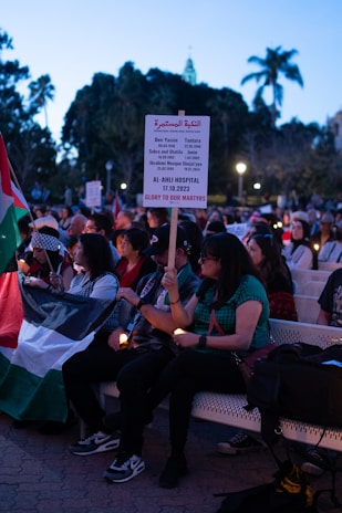 A group of people sitting on benches, some holding candles. One person is holding a sign that lists historical events and dates, along with a phrase expressing honor to martyrs. The scene takes place outdoors in a public park with trees and lamps visible in the background. Some individuals are holding a flag, and the overall setting appears to be a peaceful gathering or vigil.
