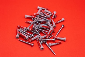 A scattered pile of metal screws and plastic wall plugs is set against a vibrant red background. The screws have a shiny silver appearance, while the wall plugs are predominantly gray with some red accents.