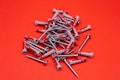 A scattered pile of metal screws and plastic wall plugs is set against a vibrant red background. The screws have a shiny silver appearance, while the wall plugs are predominantly gray with some red accents.