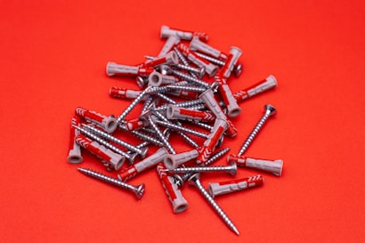 A scattered pile of metal screws and plastic wall plugs is set against a vibrant red background. The screws have a shiny silver appearance, while the wall plugs are predominantly gray with some red accents.