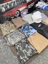 A collection of folded clothes laid out on a textured surface, including camouflage shirts and pants, denim jeans, and patterned shirts. There is also a white hat placed on top of some clothing items, and a backdrop featuring a classic red car and a scarf with 'Mickey' written on it.