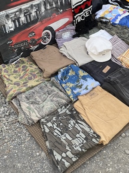 A collection of folded clothes laid out on a textured surface, including camouflage shirts and pants, denim jeans, and patterned shirts. There is also a white hat placed on top of some clothing items, and a backdrop featuring a classic red car and a scarf with 'Mickey' written on it.