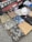 A collection of folded clothes laid out on a textured surface, including camouflage shirts and pants, denim jeans, and patterned shirts. There is also a white hat placed on top of some clothing items, and a backdrop featuring a classic red car and a scarf with 'Mickey' written on it.