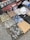 A collection of folded clothes laid out on a textured surface, including camouflage shirts and pants, denim jeans, and patterned shirts. There is also a white hat placed on top of some clothing items, and a backdrop featuring a classic red car and a scarf with 'Mickey' written on it.