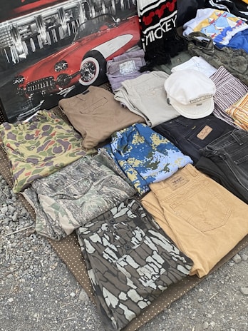 A collection of folded clothes laid out on a textured surface, including camouflage shirts and pants, denim jeans, and patterned shirts. There is also a white hat placed on top of some clothing items, and a backdrop featuring a classic red car and a scarf with 'Mickey' written on it.
