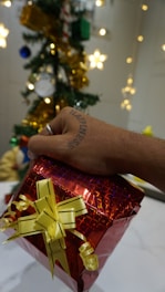 A hand with a tattoo that reads 'Happiness' is holding a red gift box wrapped with yellow ribbon. In the background, there is a decorated Christmas tree with ornaments like stars, gold garlands, and baubles.