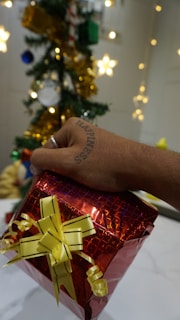 A hand with a tattoo that reads 'Happiness' is holding a red gift box wrapped with yellow ribbon. In the background, there is a decorated Christmas tree with ornaments like stars, gold garlands, and baubles.