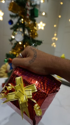 A hand with a tattoo that reads 'Happiness' is holding a red gift box wrapped with yellow ribbon. In the background, there is a decorated Christmas tree with ornaments like stars, gold garlands, and baubles.
