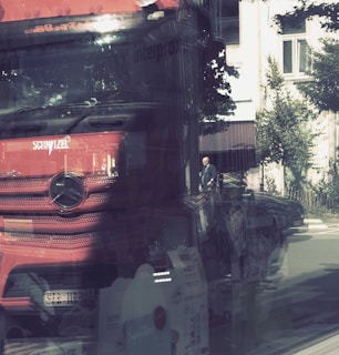 A large red truck with a prominent grille and logo is reflected in a glass window. The reflection merges with the view of an outdoor street scene where a person in dark clothing is walking on the sidewalk. Nearby trees and a building with white walls and windows are visible in the background. The image has a layered, abstract quality due to the reflections.