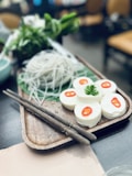 A wooden tray displays sliced white tofu garnished with red chili slices and a parsley leaf. Beside it, delicate bundles of rice noodles are placed on a green leaf. Two wooden chopsticks rest on the tray. In the background, leafy greens are slightly blurred.