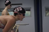 Two swimmers are preparing to race, wearing black swim caps with red and white lettering. The foreground swimmer is leaning forward with focus, while the background swimmer stands upright, both wearing goggles.