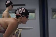 Two swimmers are preparing to race, wearing black swim caps with red and white lettering. The foreground swimmer is leaning forward with focus, while the background swimmer stands upright, both wearing goggles.