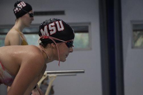 Two swimmers are preparing to race, wearing black swim caps with red and white lettering. The foreground swimmer is leaning forward with focus, while the background swimmer stands upright, both wearing goggles.