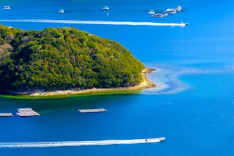 A lush, green island surrounded by clear blue water. Several boats are navigating the water with visible wakes, and there are floating docks or platforms near the island.