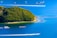 A lush, green island surrounded by clear blue water. Several boats are navigating the water with visible wakes, and there are floating docks or platforms near the island.