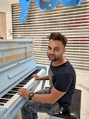 A person is seated at a light blue upright piano, playing the keys with both hands. The individual has short hair and is wearing a black t-shirt and jeans. Behind the person, there is a closed shop with a silver and white shutter and a large blue sign with letters in a stylized font.