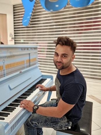 A person is seated at a light blue upright piano, playing the keys with both hands. The individual has short hair and is wearing a black t-shirt and jeans. Behind the person, there is a closed shop with a silver and white shutter and a large blue sign with letters in a stylized font.