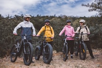 Four people equipped with helmets and colorful jackets stand with their fat-tire bicycles on a grassy area surrounded by greenery. The sky is overcast, adding a moody atmosphere to the scene.