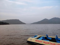 A serene lake with calm waters reflecting the overcast sky is flanked by rolling hills in the distance. A paddle boat with the words 'Wonder Mountain' is partially visible in the foreground, featuring bright blue seats.